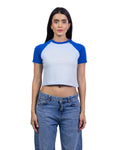 A woman modeling a short-sleeve crop top with a white body and royal blue raglan sleeves, paired with blue jeans, against a plain white background.