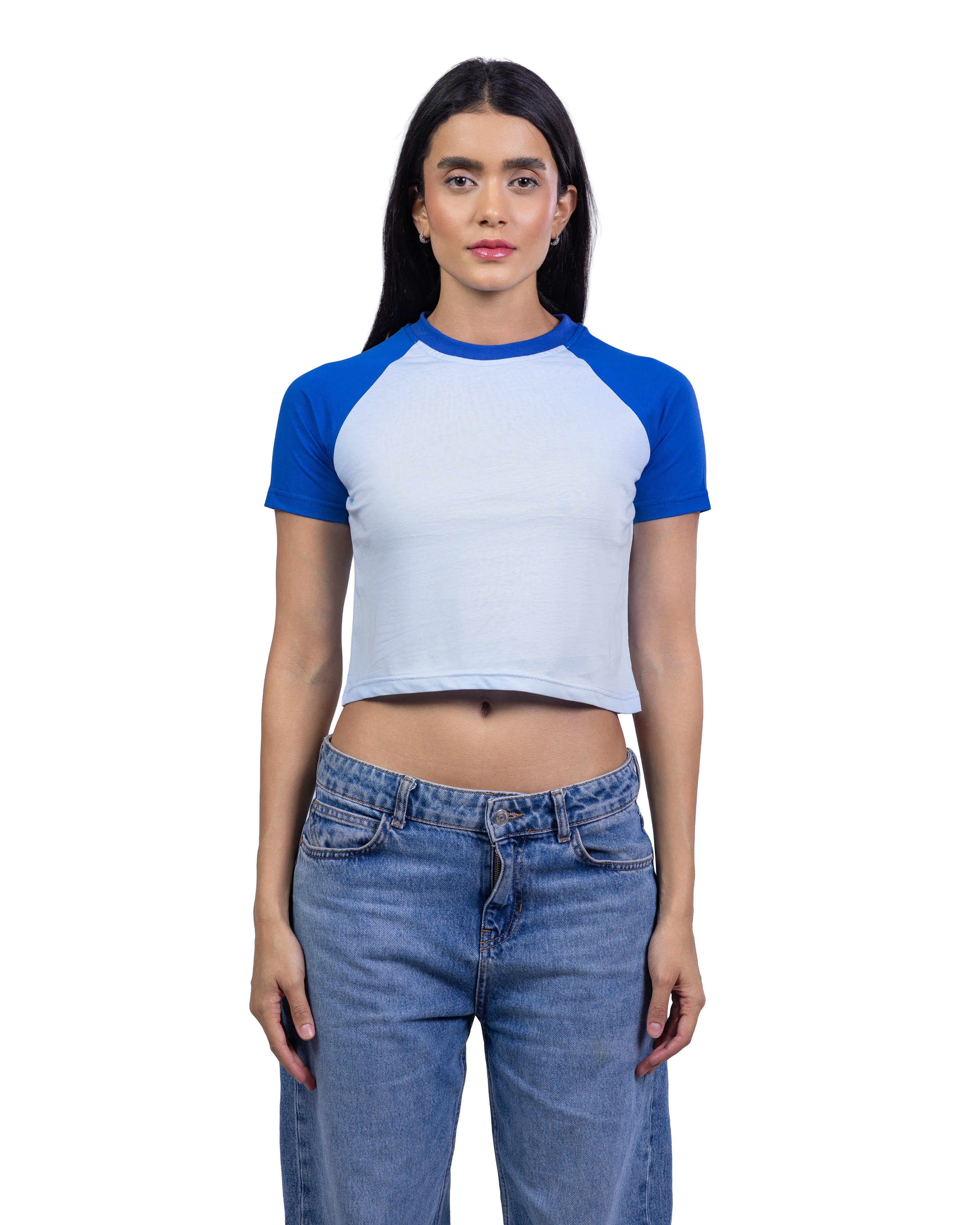 A woman modeling a short-sleeve crop top with a white body and royal blue raglan sleeves, paired with blue jeans, against a plain white background.