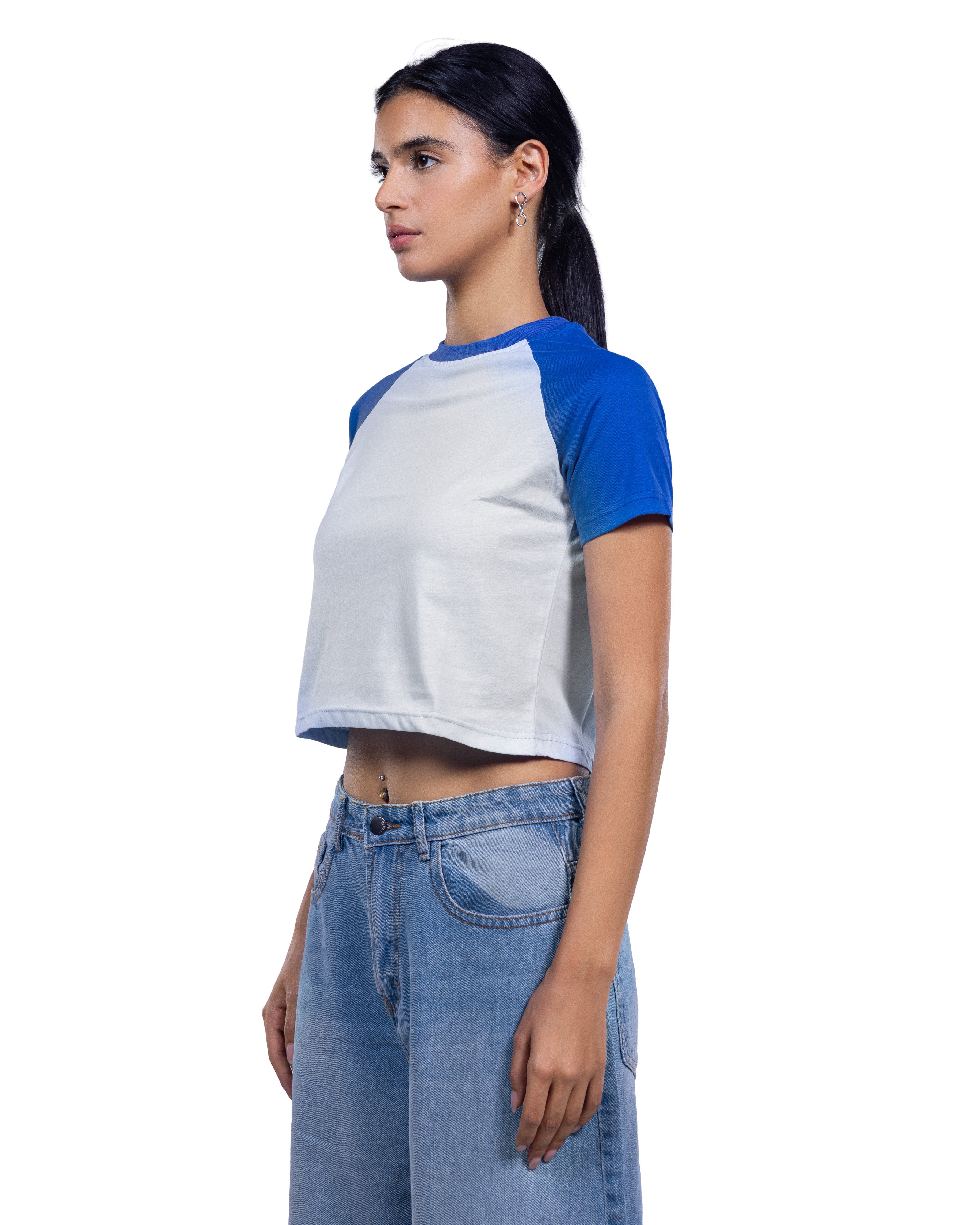 A woman wearing a short-sleeve raglan crop top with a white body and blue sleeves, paired with light blue jeans, shown in profile against a plain white background.
