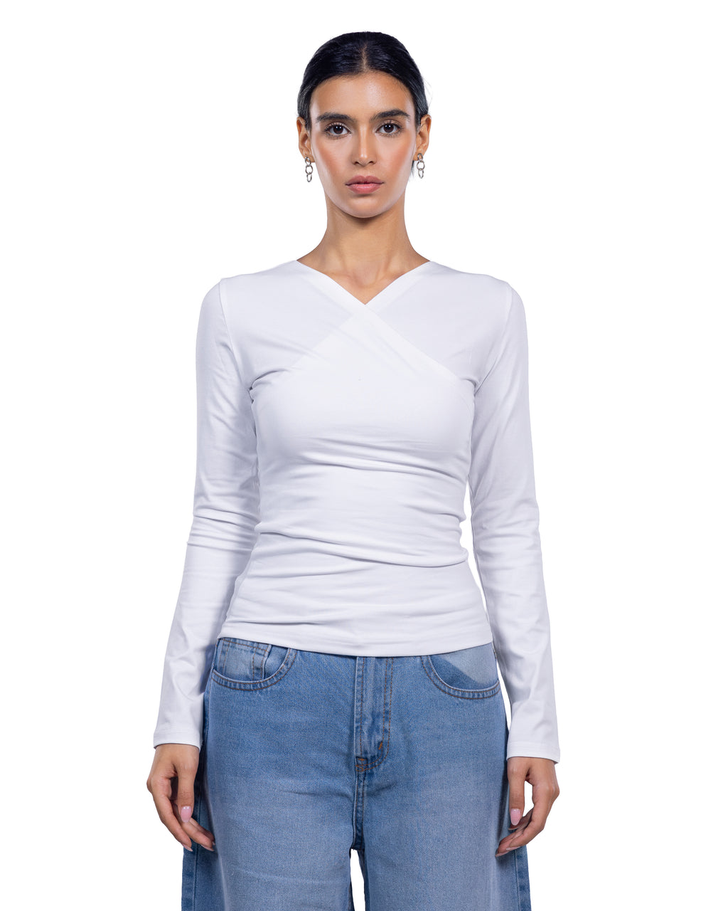 A woman modeling a fitted white long-sleeve top with a crossover V-neck design, paired with light blue jeans, against a plain white background.