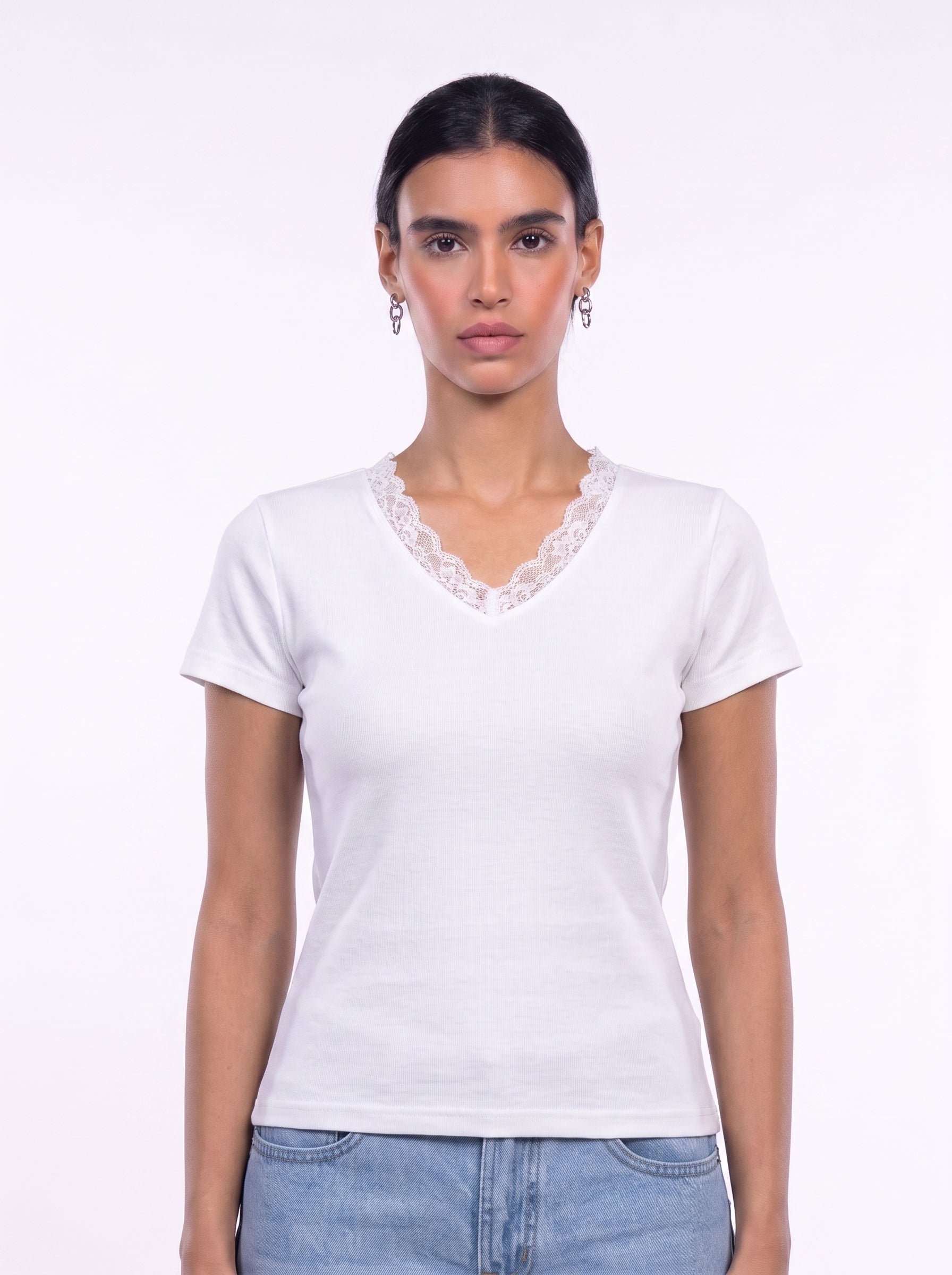 A woman modeling a white short-sleeve V-neck t-shirt with a delicate lace trim along the neckline, paired with light blue jeans against a plain white studio background.