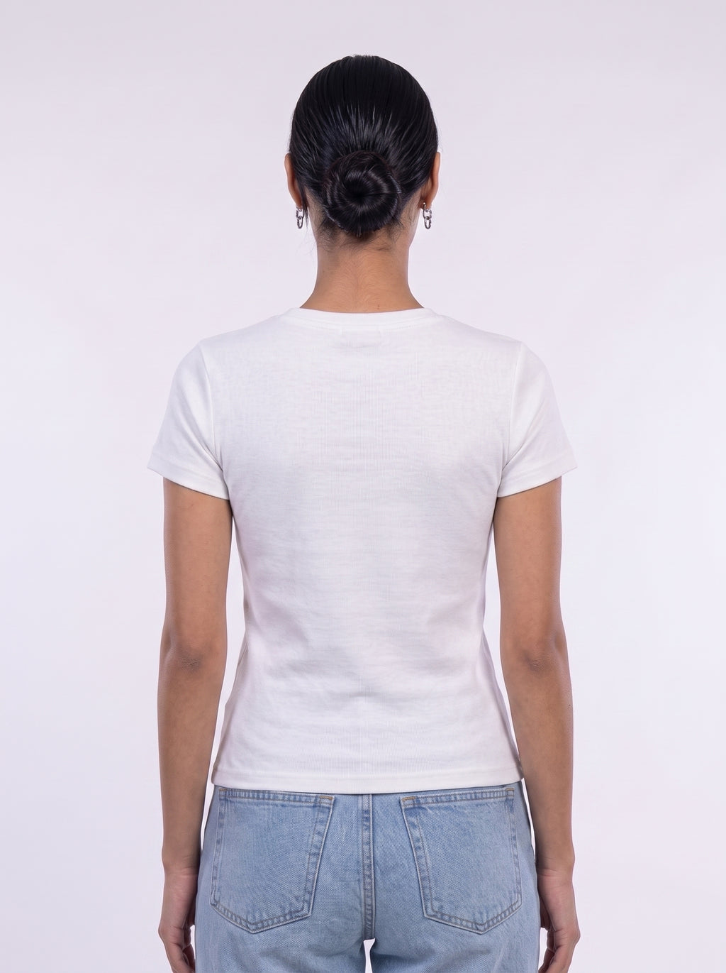 A woman viewed from behind wearing a plain white short-sleeve crewneck t-shirt and light blue jeans against a light gray studio background.