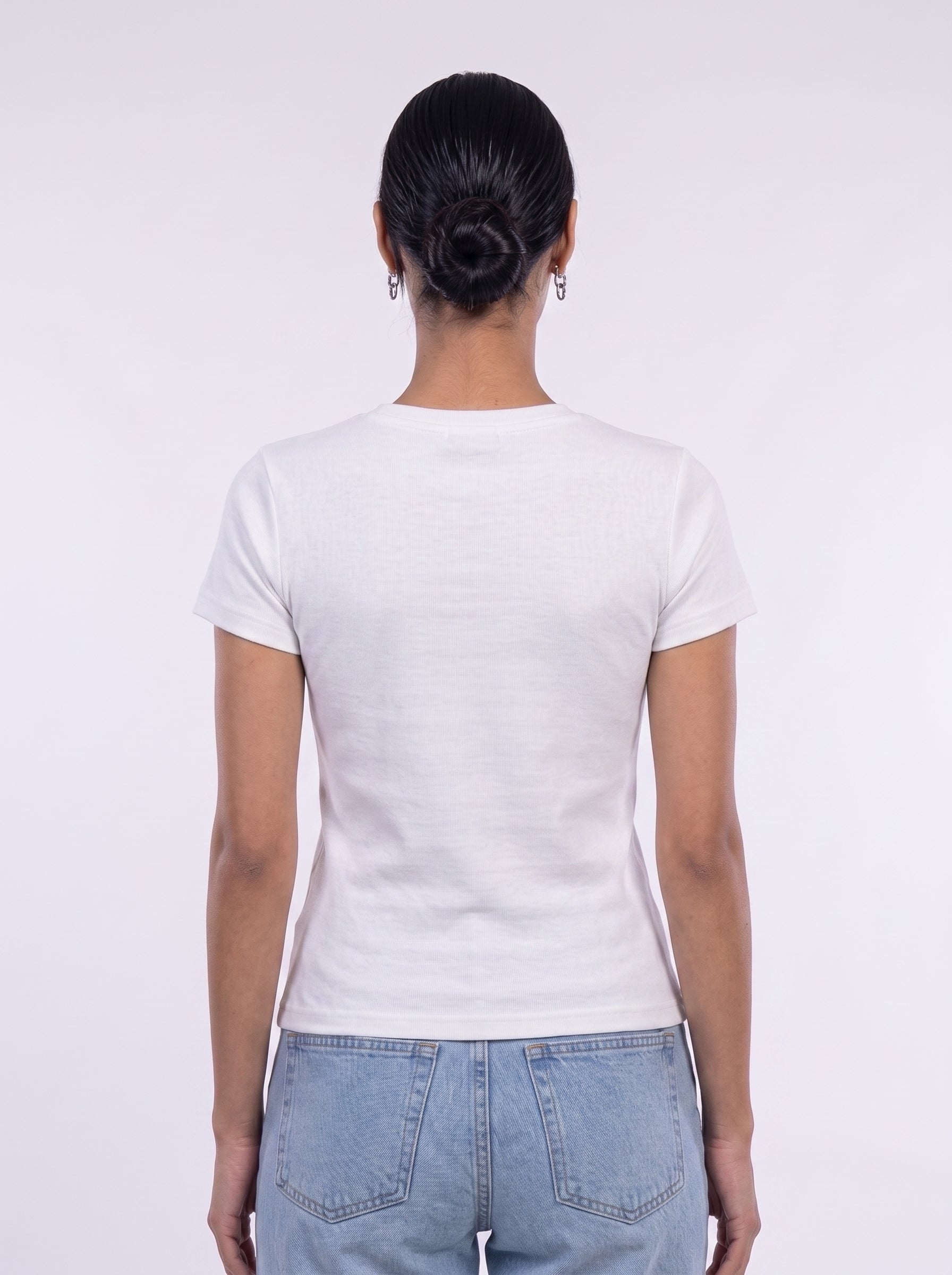 A woman viewed from behind wearing a plain white short-sleeve crewneck t-shirt and light blue jeans against a light gray studio background.