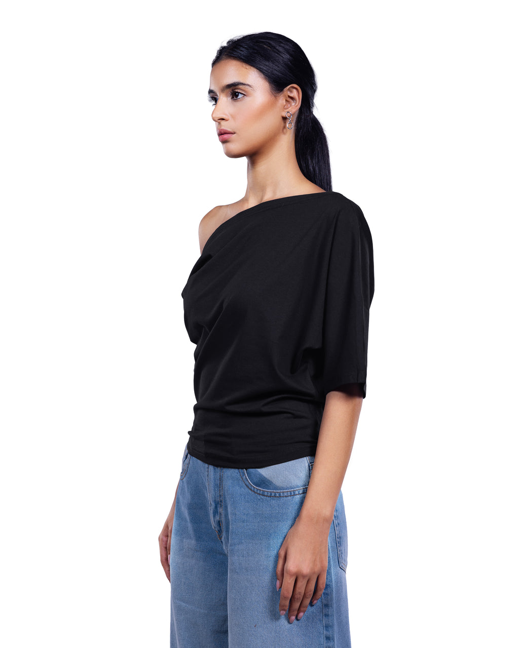 A woman modeling a black one-shoulder draped top with elbow-length sleeves, paired with light blue jeans, against a plain white background.