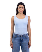 A woman modeling a light blue sleeveless bodysuit or tank top paired with blue jeans, shot against a solid white background.