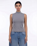 A woman modeling a sleeveless gray mock neck top paired with blue jeans against a plain white studio background.