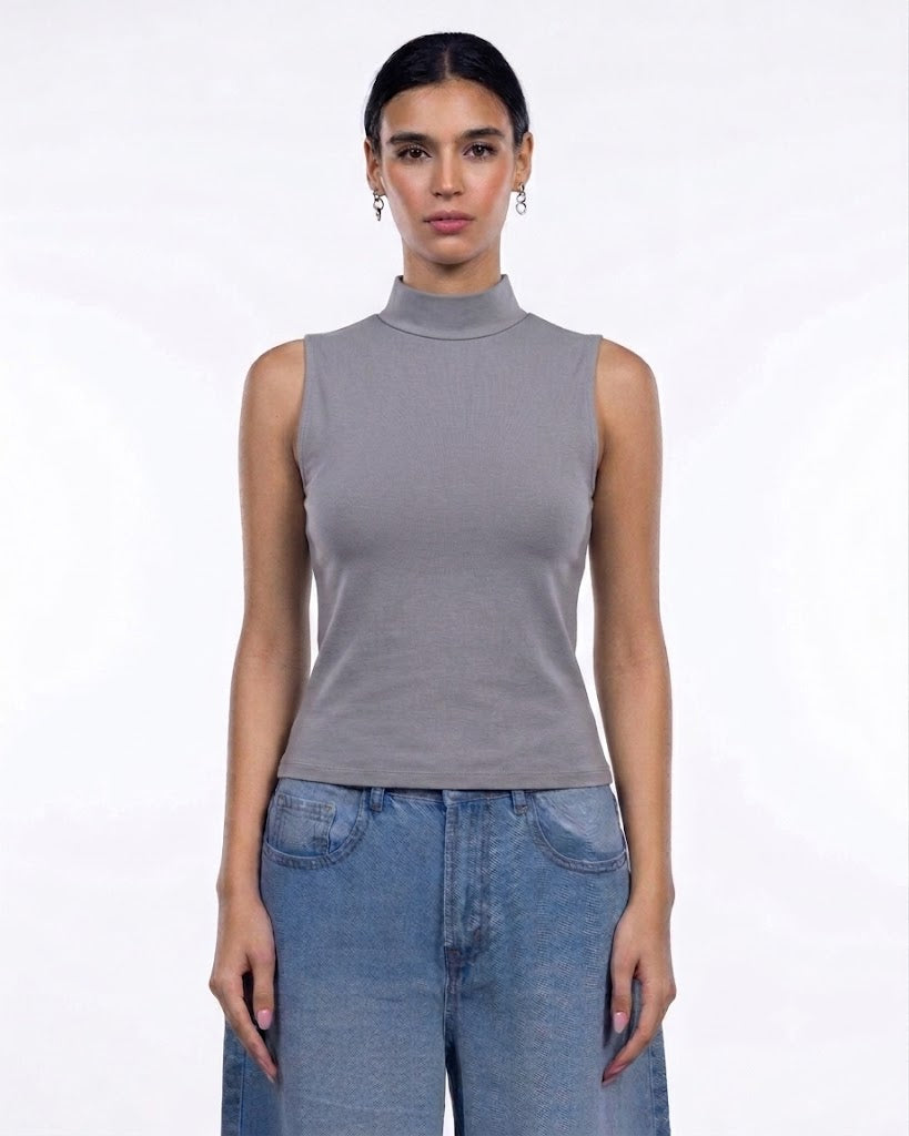 A woman modeling a sleeveless gray mock neck top paired with blue jeans against a plain white studio background.
