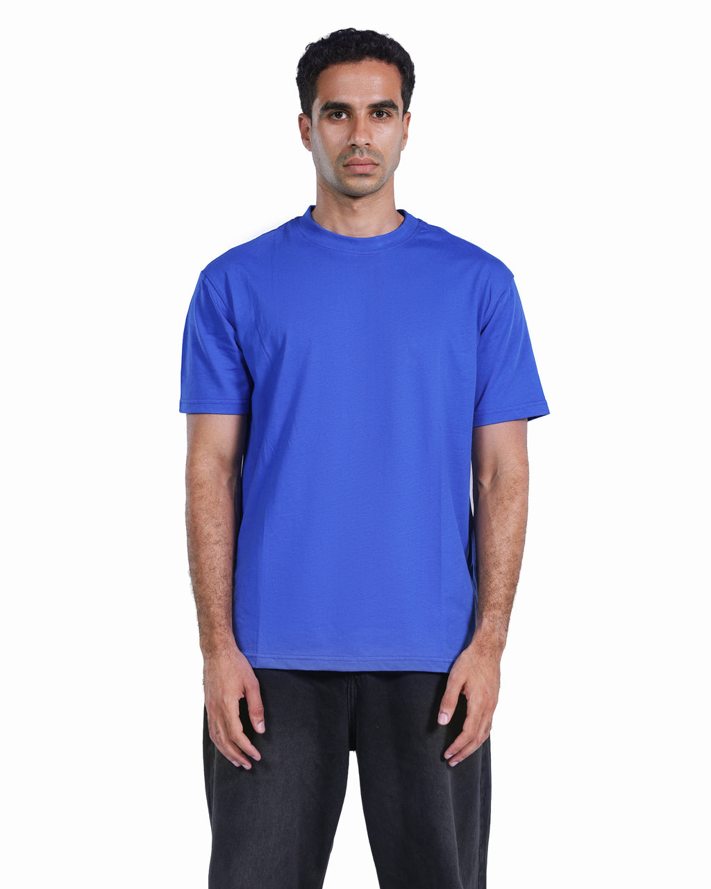 A man wearing a solid royal blue short-sleeve crewneck t-shirt and dark gray pants, standing against a plain white background.