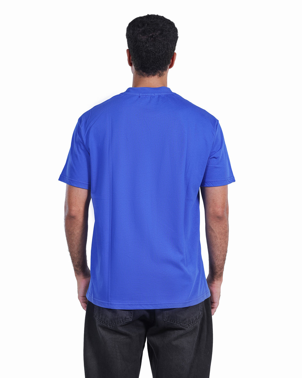 A man viewed from behind wearing a solid royal blue short-sleeve t-shirt and dark gray pants, set against a plain white background.