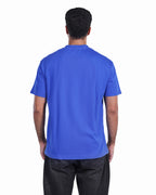 A man viewed from behind wearing a solid royal blue short-sleeve t-shirt and dark gray pants, set against a plain white background.