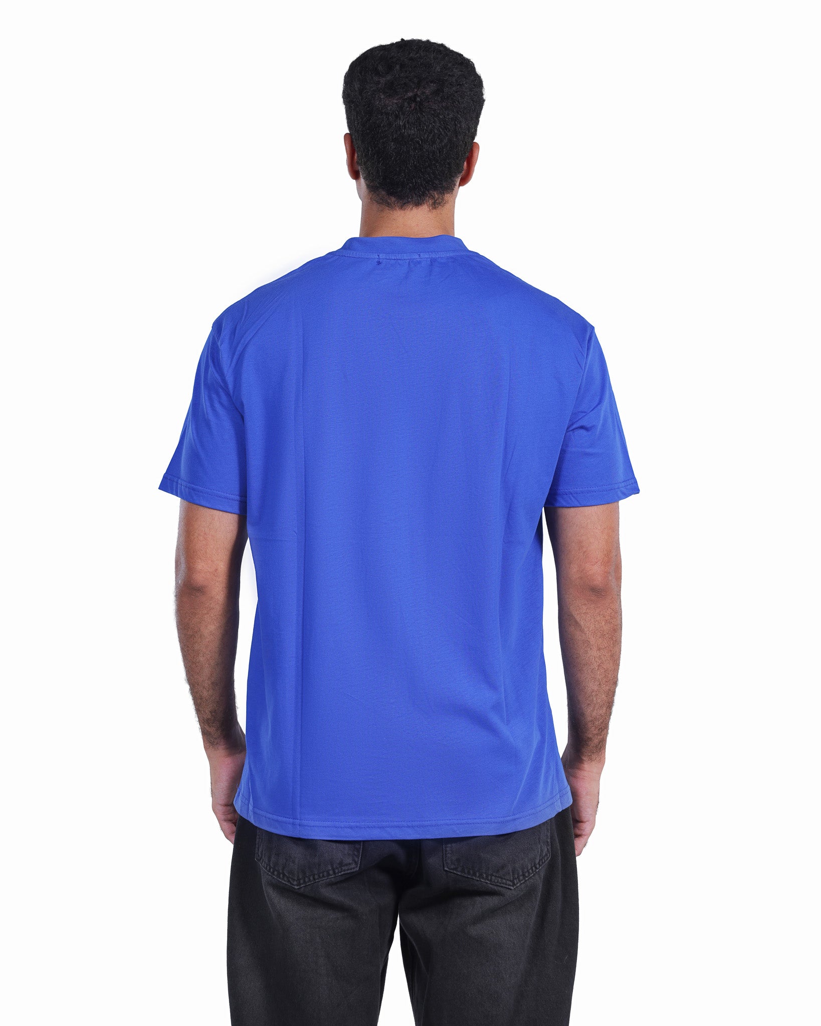 A man viewed from behind wearing a solid royal blue short-sleeve t-shirt and dark gray pants, set against a plain white background.