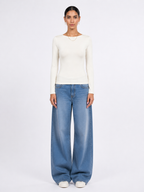 A model stands against a plain white studio background wearing a fitted off-white long-sleeve top, wide-leg light blue jeans, and white shoes. [model:1]