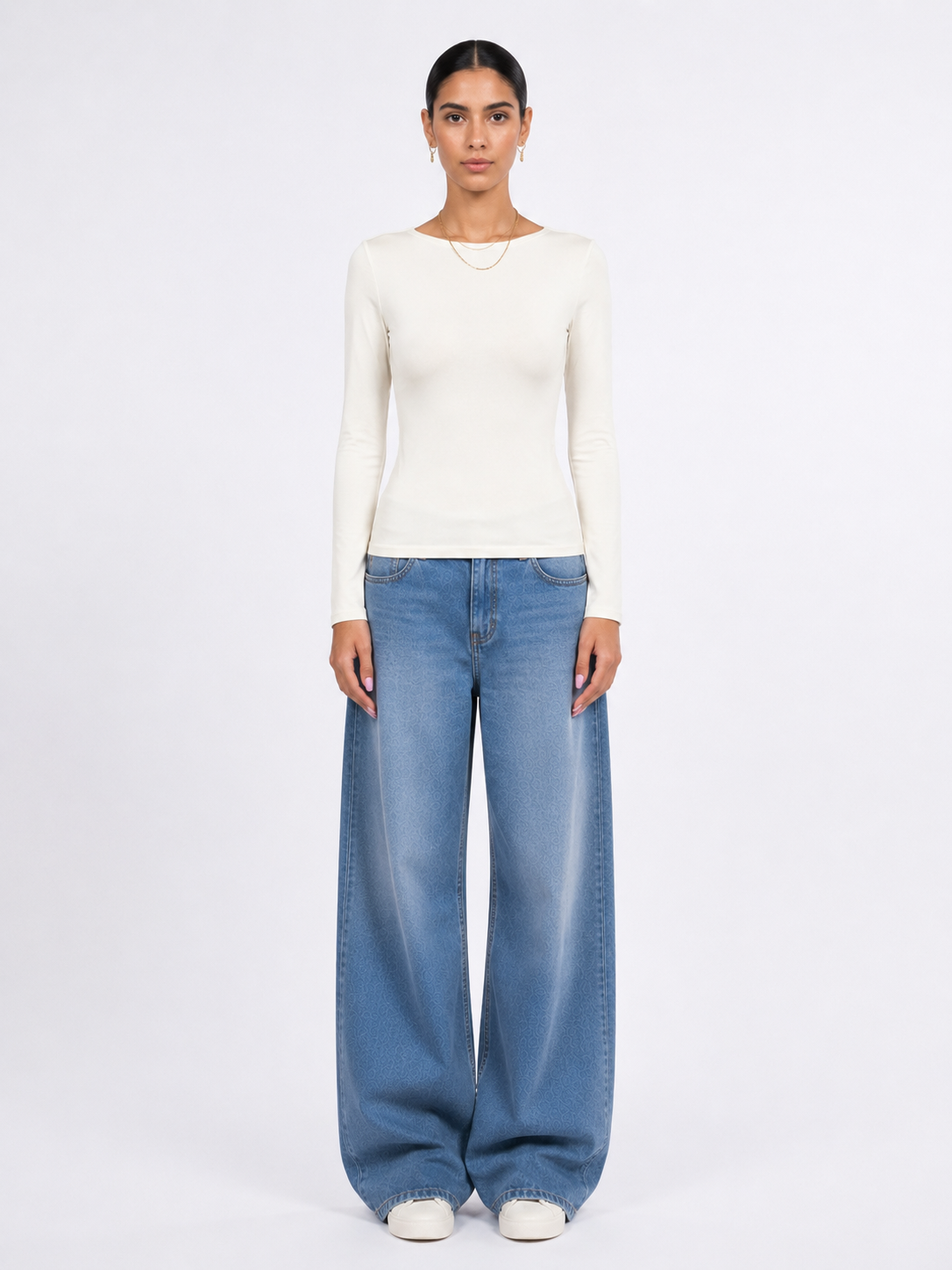 A model stands against a plain white studio background wearing a fitted off-white long-sleeve top, wide-leg light blue jeans, and white shoes.