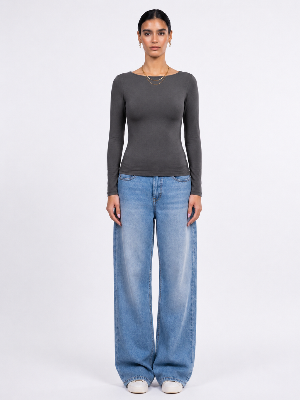 A model stands against a light gray studio background wearing a dark gray long-sleeve boat-neck top and wide-leg light blue jeans.