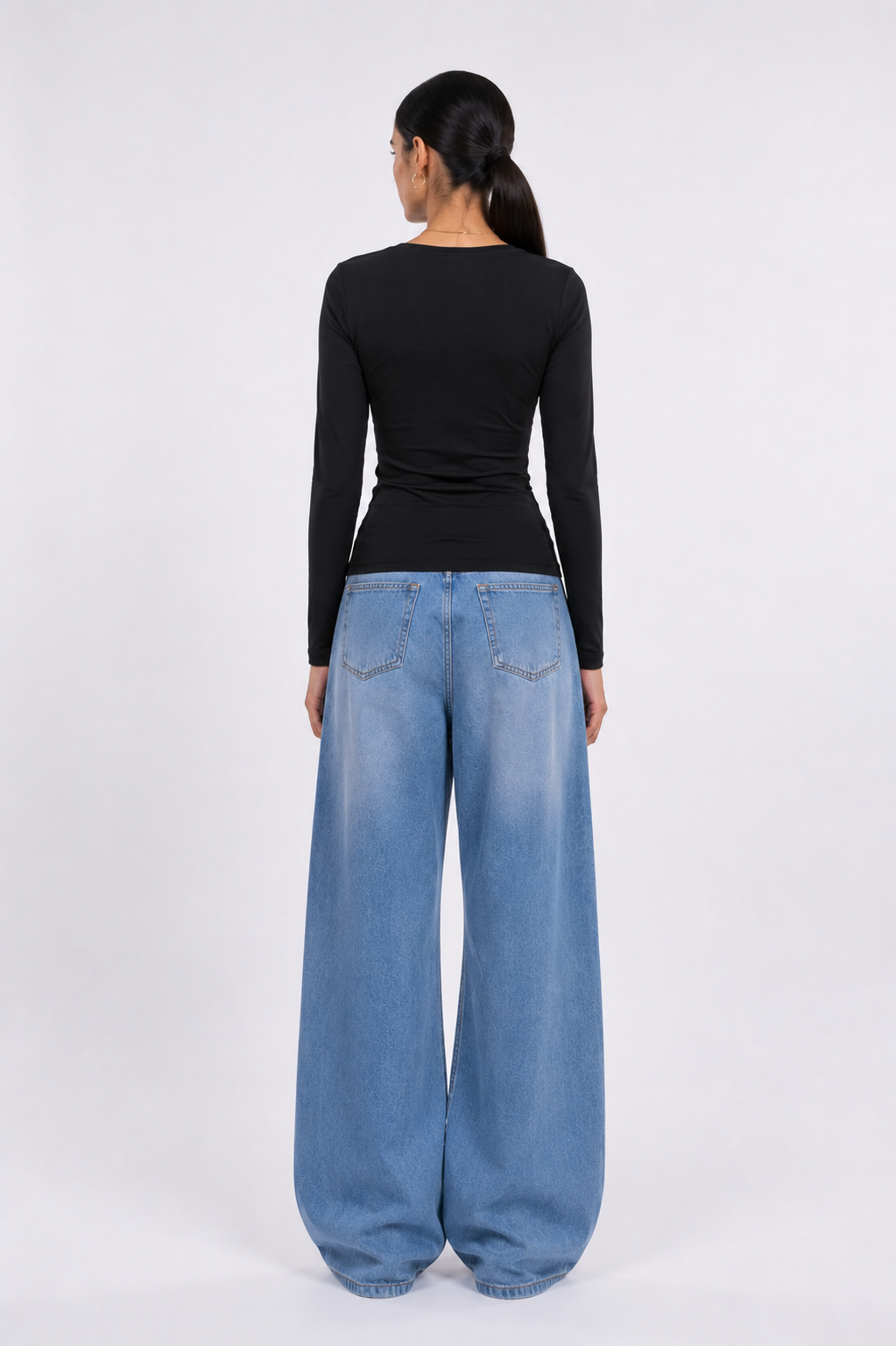 A woman shown from the back wearing a fitted black long-sleeve top and wide-leg light blue denim jeans against a plain light gray studio background.