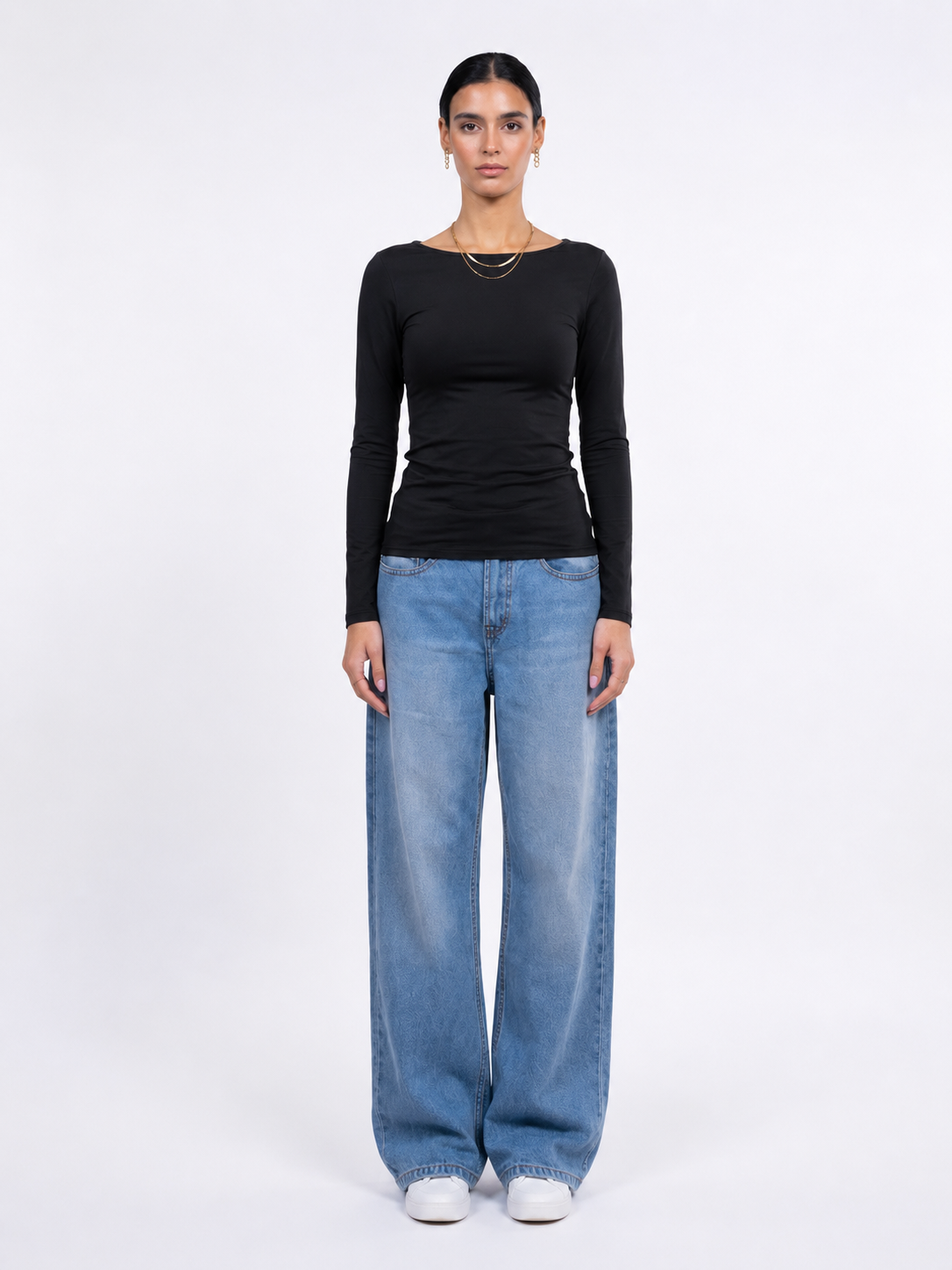 A model stands against a plain white studio background wearing a fitted black long-sleeve top, wide-leg light blue jeans, and white sneakers.