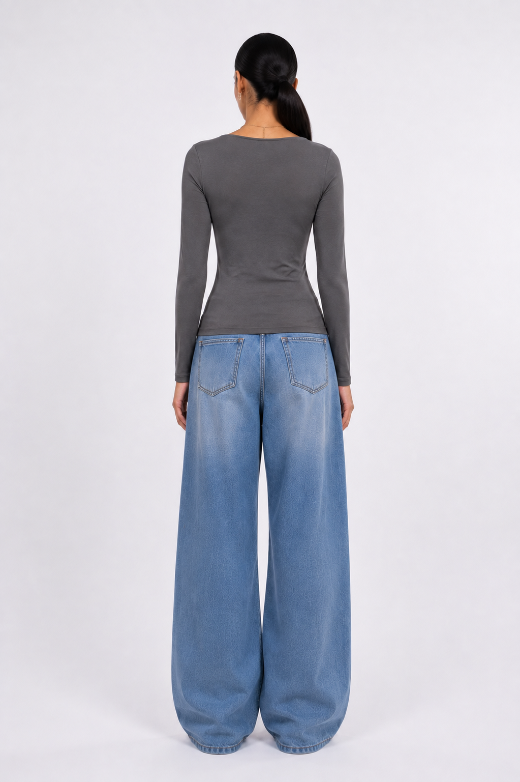 A woman shown from the back wearing a dark gray long-sleeve top and wide-leg light blue denim jeans against a plain light gray studio background.