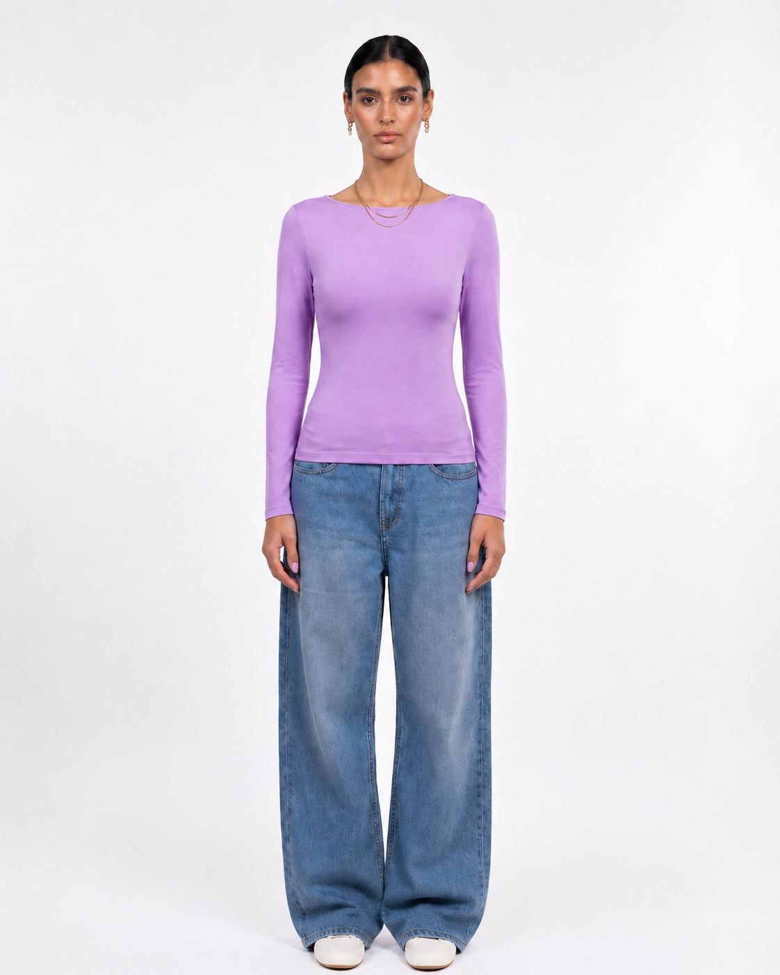 A model stands against a plain white studio background wearing a fitted lavender long-sleeve top and wide-leg blue jeans.