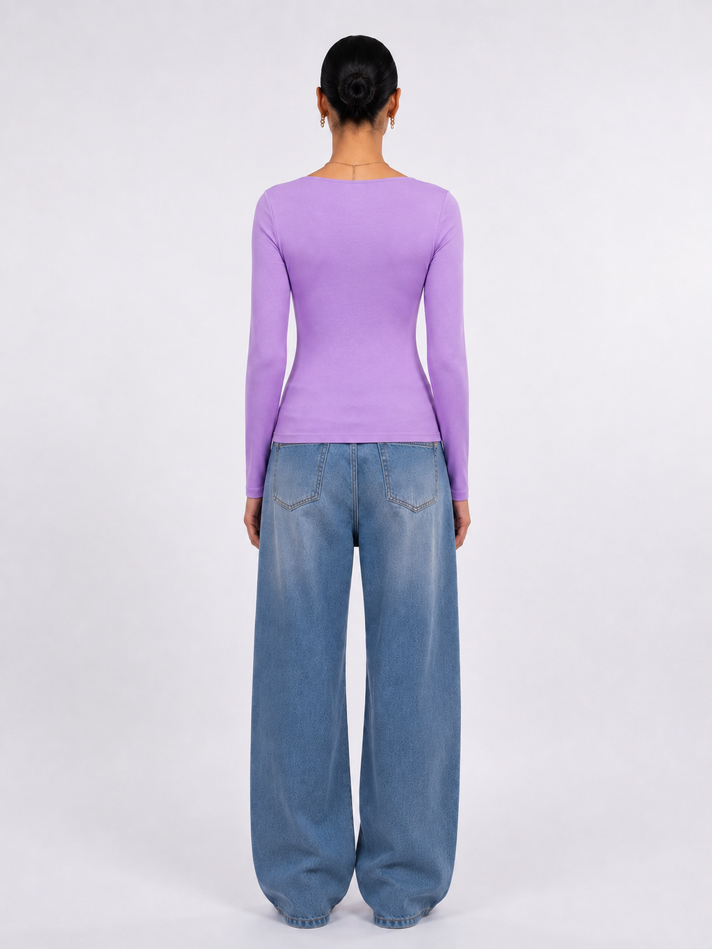A model viewed from the back wearing a fitted lavender long-sleeve top and wide-leg blue jeans against a plain light gray studio background.
