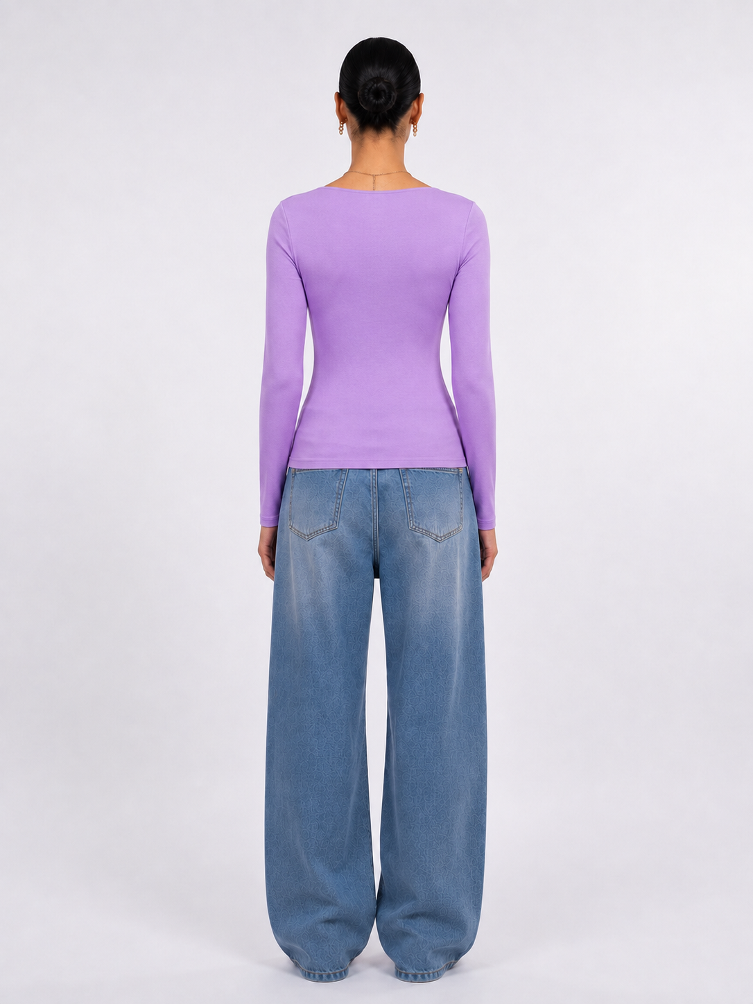 A model viewed from the back wearing a fitted lavender long-sleeve top and wide-leg blue jeans against a plain light gray studio background.