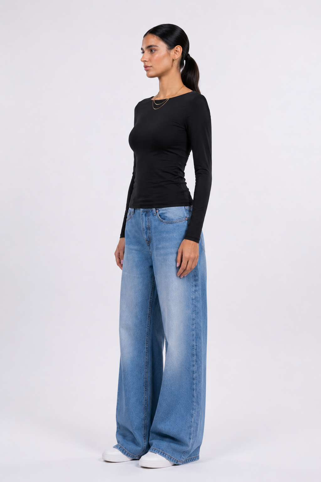 A woman modeling a black long-sleeve top and wide-leg light blue jeans against a plain light gray studio background.