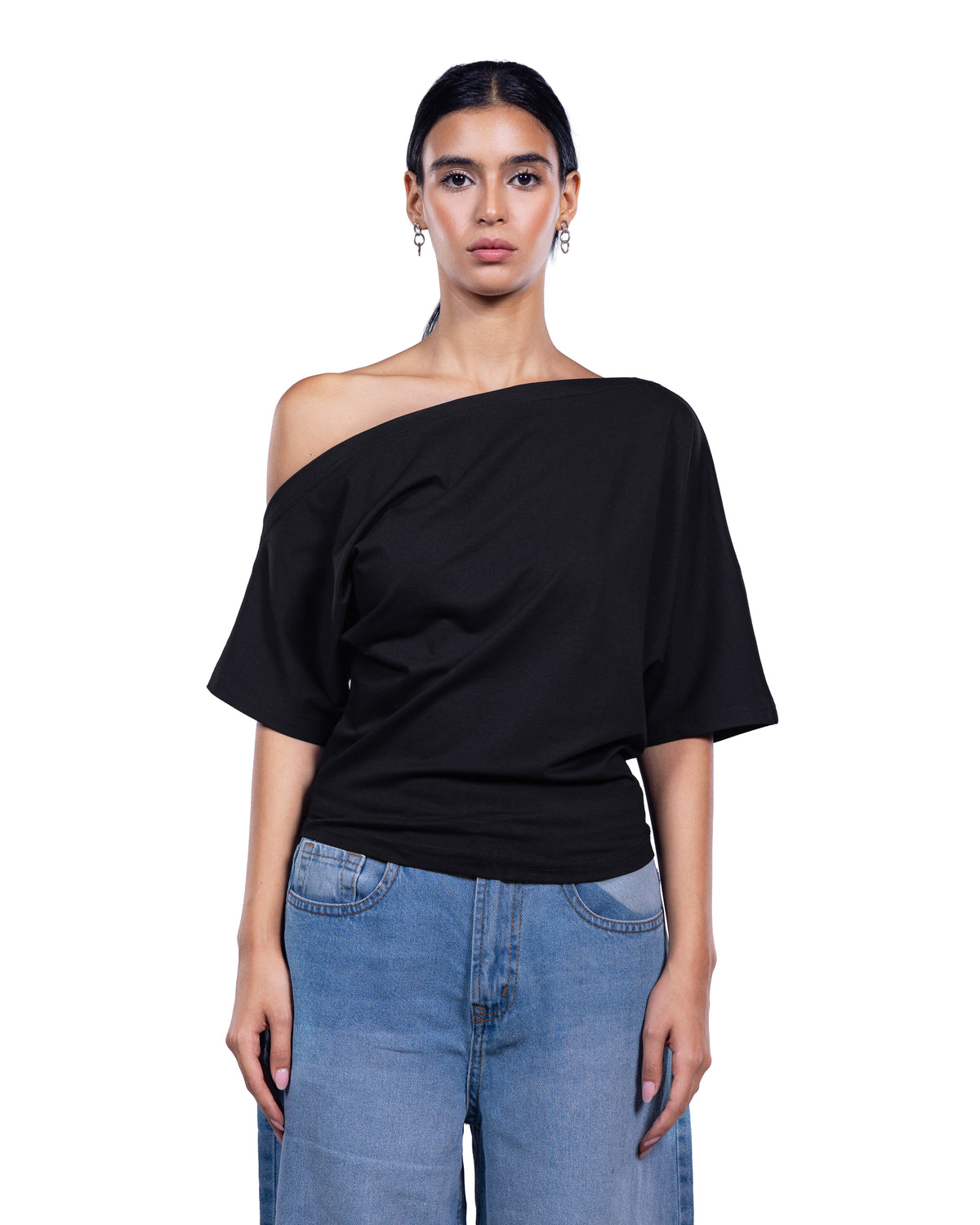A woman modeling a black off-the-shoulder short-sleeve top paired with light blue jeans, shot against a plain white background.